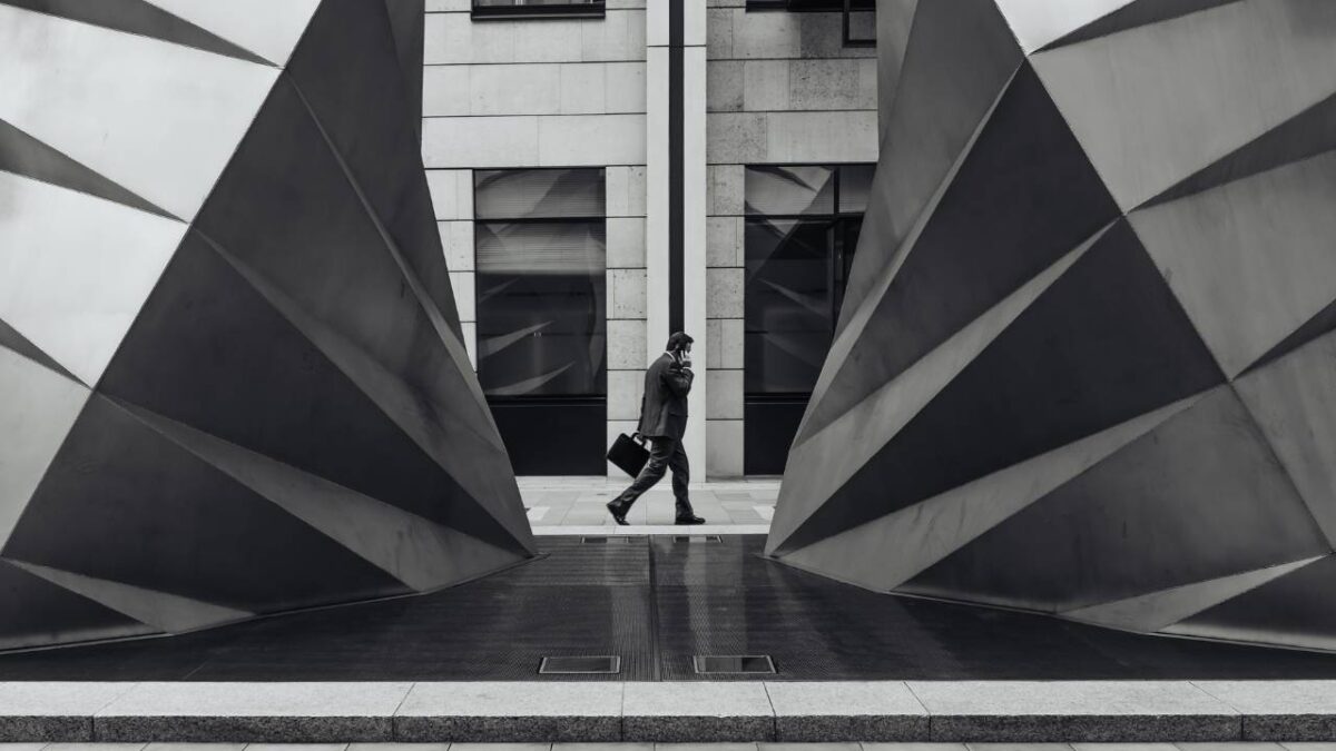 A black and white photo of a businessman walking behind a modern sculpture